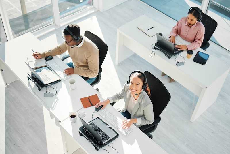 A high-angle view of a bright modern office showing three professionals working at white desks with computers and headsets in a well-lit, minimalist workspace